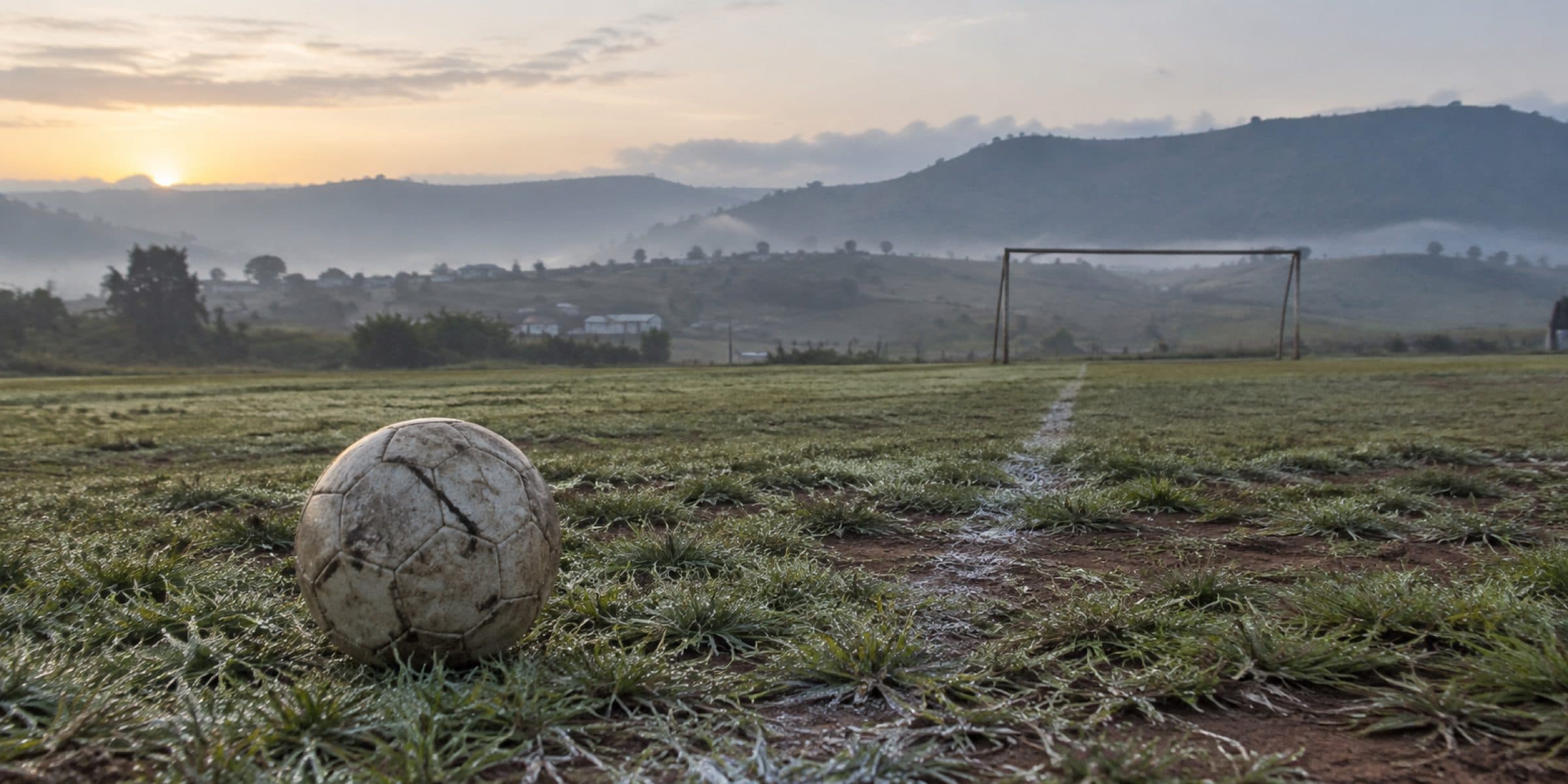 A worn match ball on a dew-covered pitch at dawn, goalposts and misted hills beyond